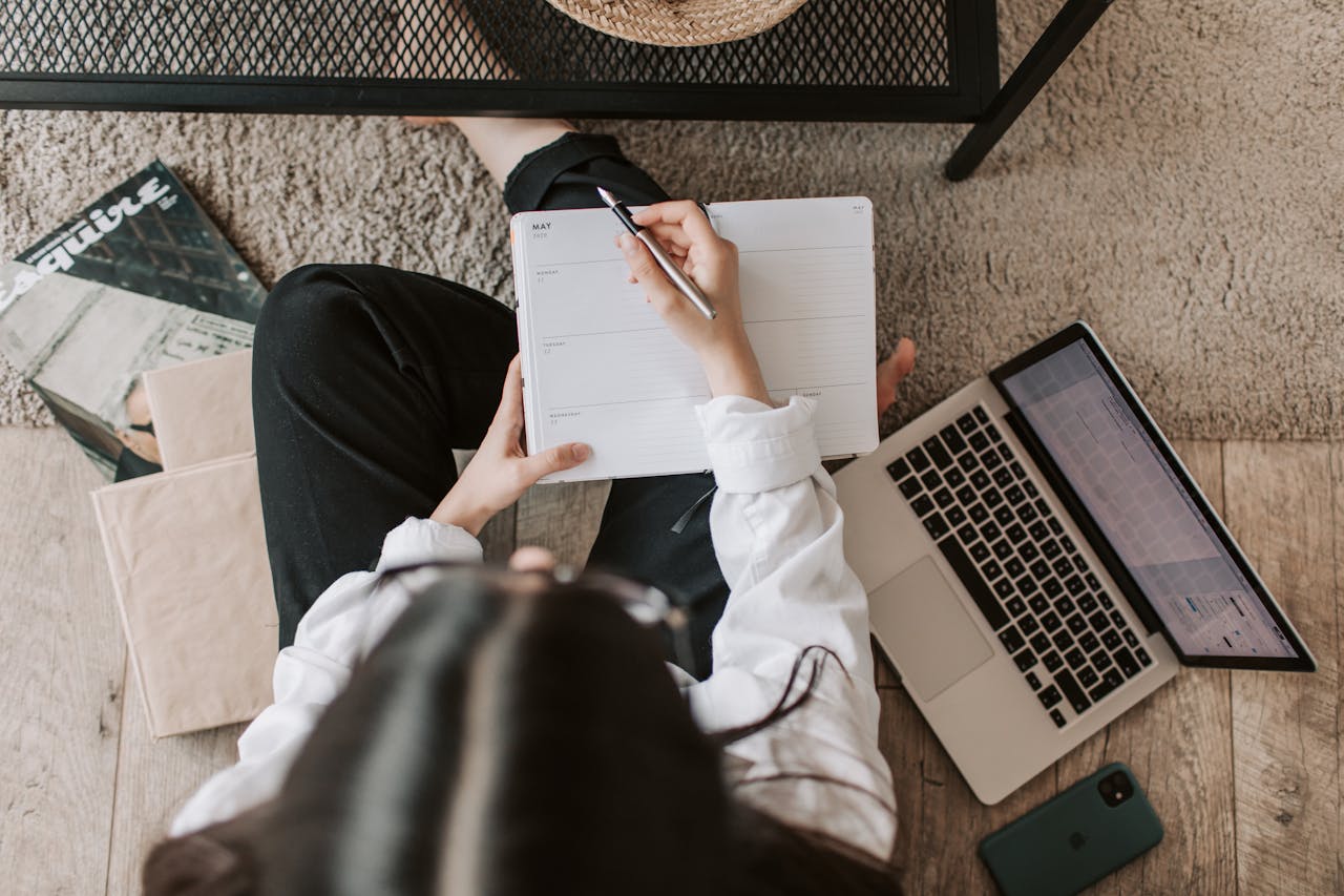 Services-03 Top view of anonymous woman in casual wear sitting on floor with laptop and smartphone and creating plan on notebook while resting during break in modern living room